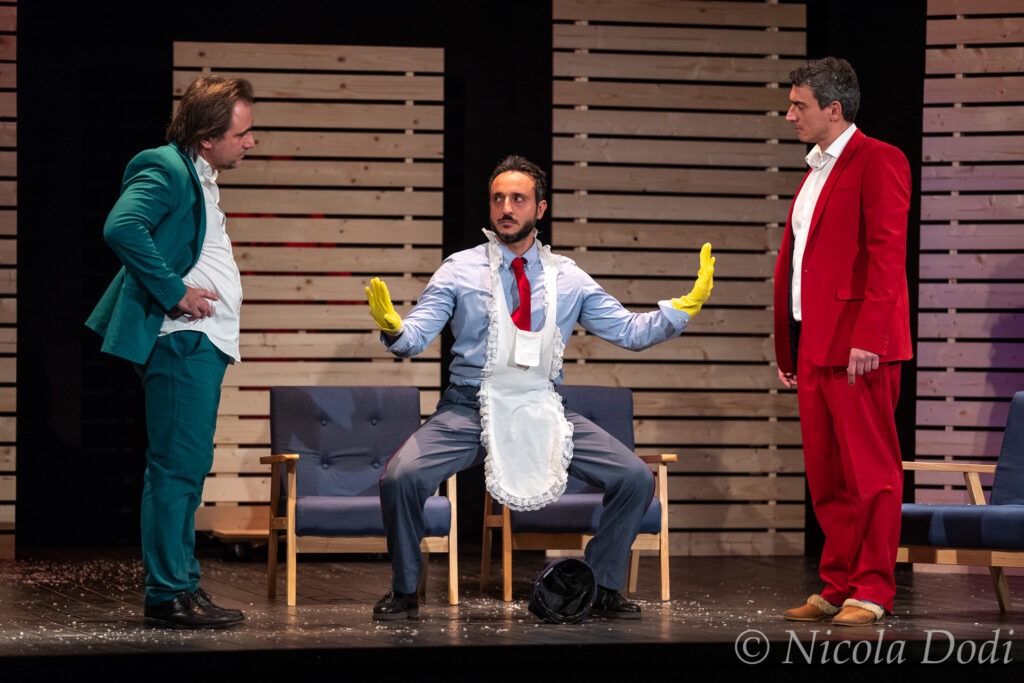 Scena della commedia Sai come son fatte le donne di On Art a Brescello: tre attori sul palco, uno con grembiule e guanti gialli, tra situazioni comiche ed equivoci.